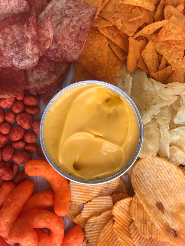 A container of cheese dip surrounded by potato chips, nacho chips and cheese puffs
